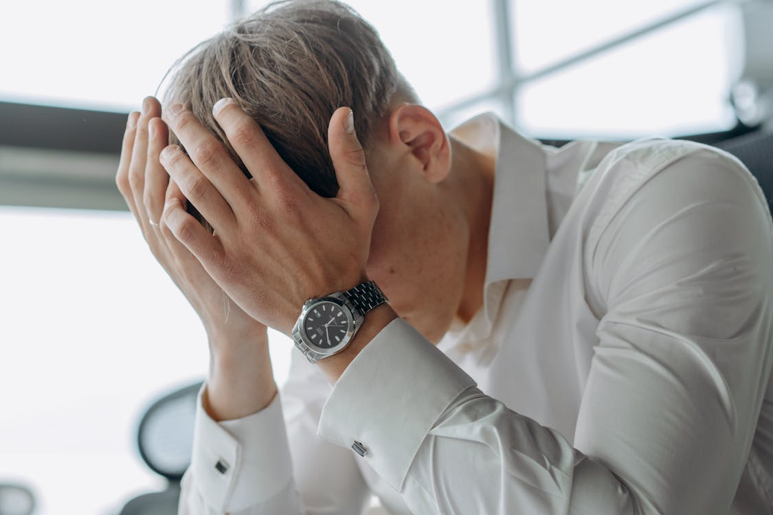 A man in a white shirt sits with his head in his hands, appearing stressed or overwhelmed in an office setting.