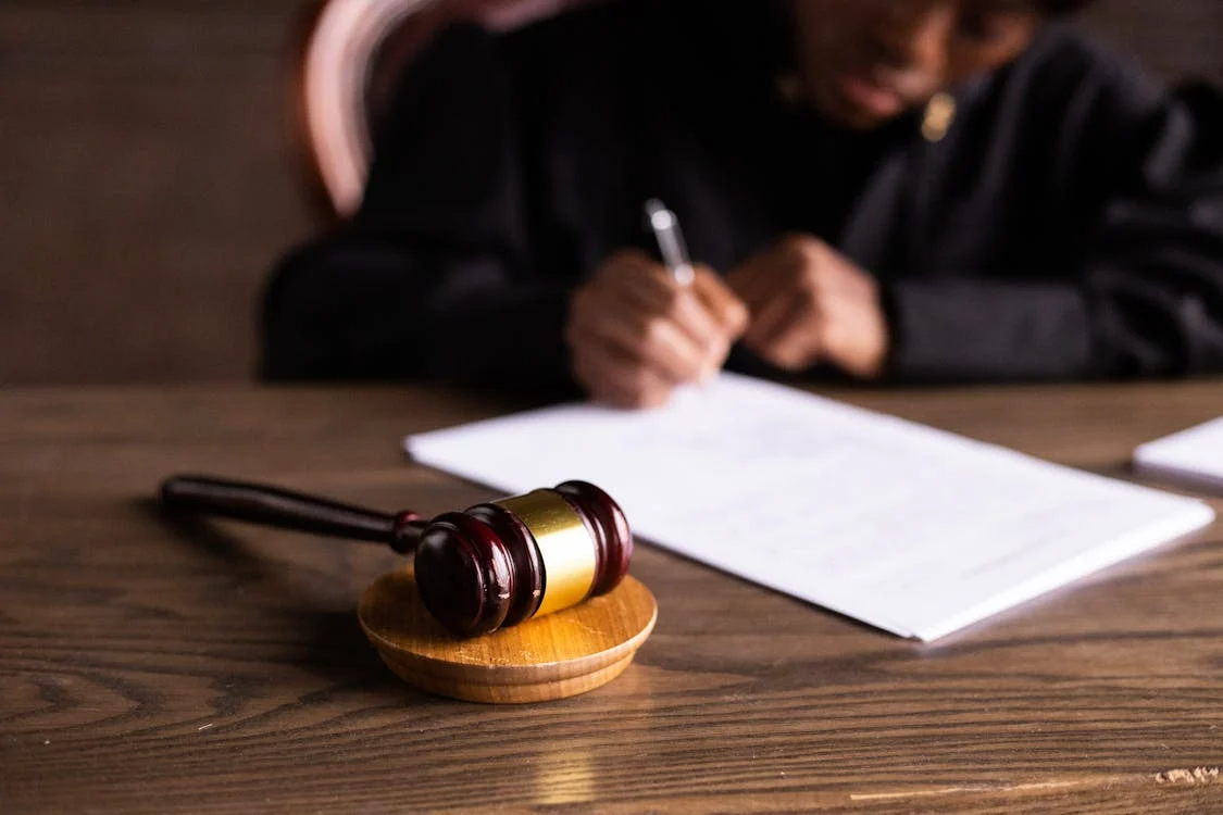 A judge’s gavel rests on a wooden table in the foreground while a person writes on a document in the background.