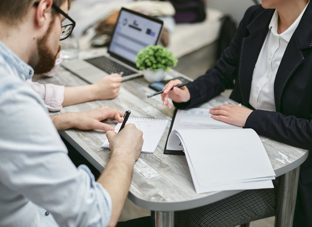 Two professionals sit at a desk discussing documents and taking notes during a business meeting.