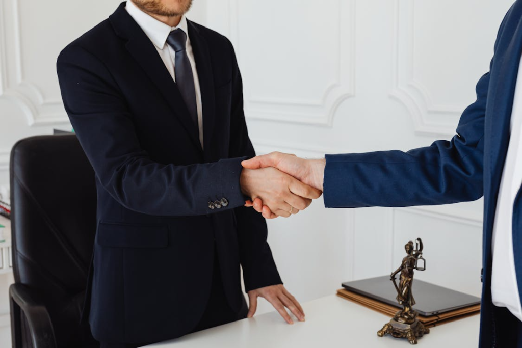 Two business professionals in suits shake hands across a desk, with a small justice statue visible nearby.