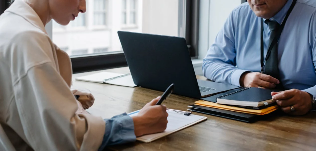 Two people sit across a desk in a professional setting, discussing or reviewing documents beside a laptop.