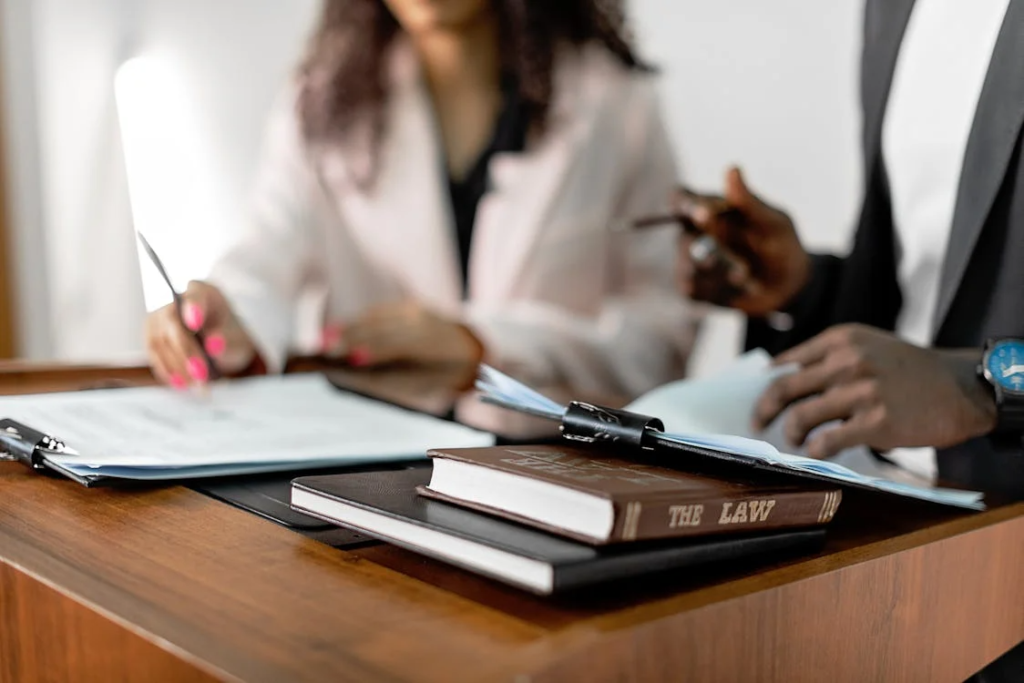 Two professionals review and sign legal documents at a desk with law books and files.