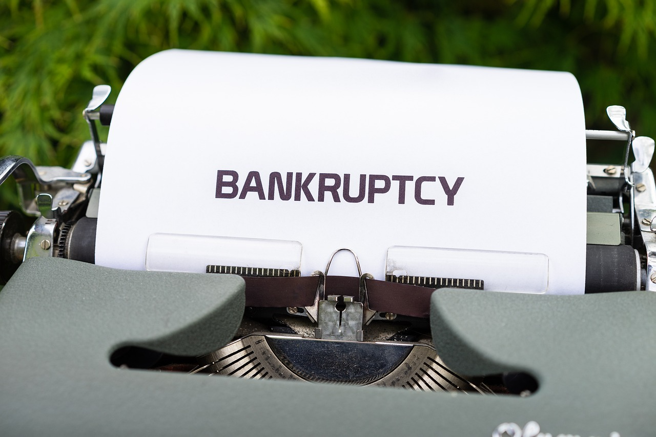  A sheet of paper reading “BANKRUPTCY” is typed into a vintage typewriter against a green outdoor background.