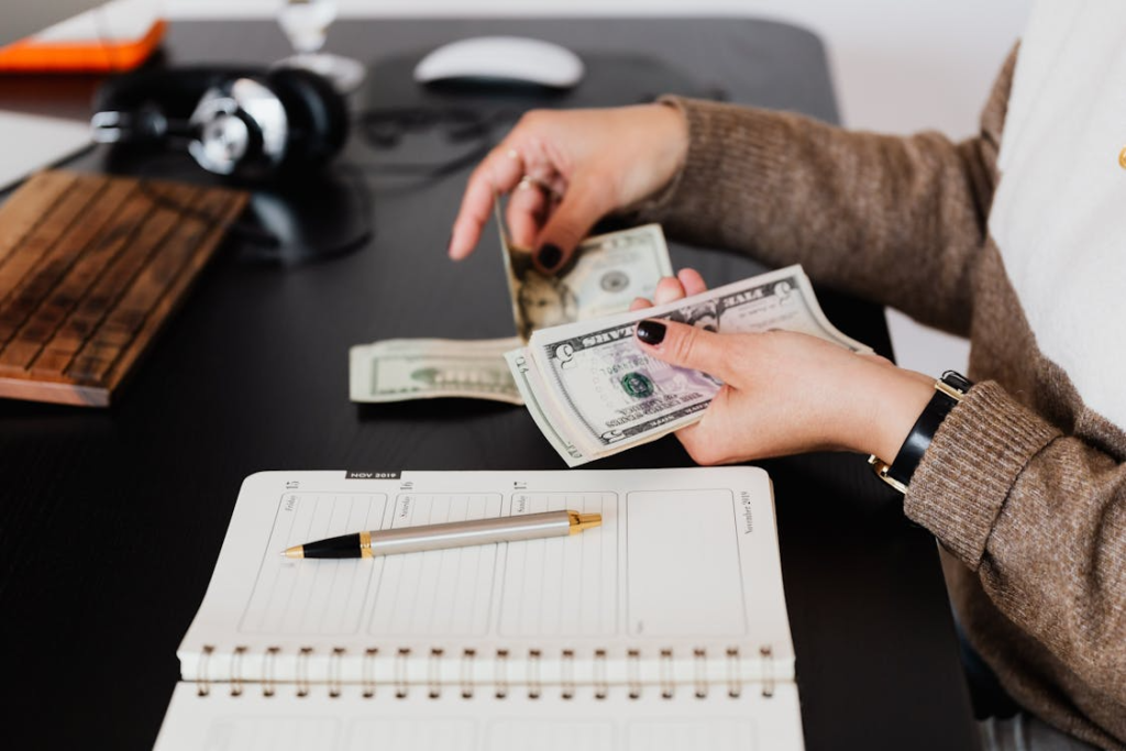 A person counts paper money at a desk with a planner and pen, suggesting budgeting or financial planning.
