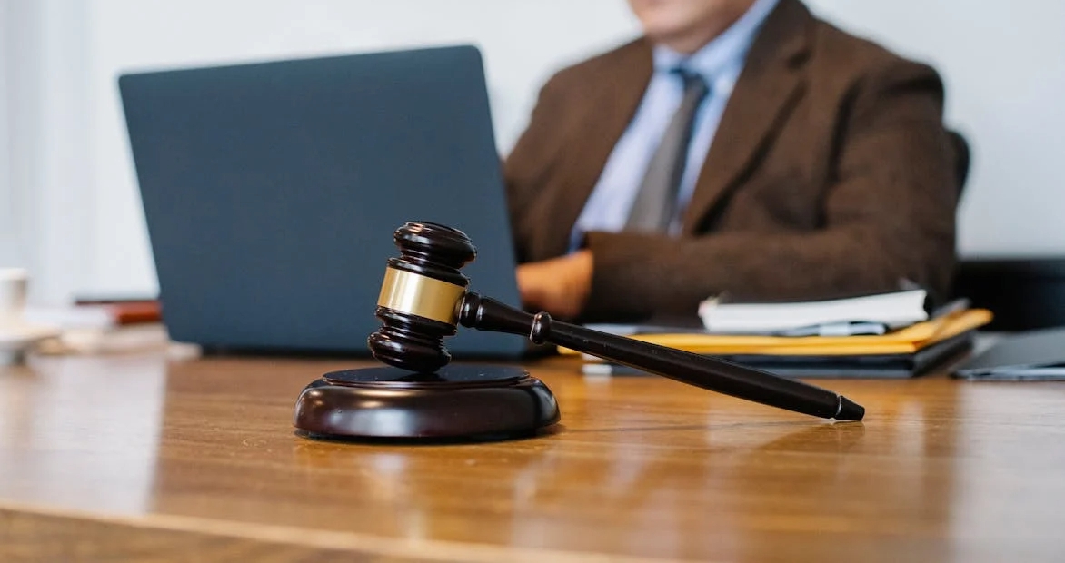 A judge’s gavel rests on a desk in the foreground while a professional works on a laptop in the background.