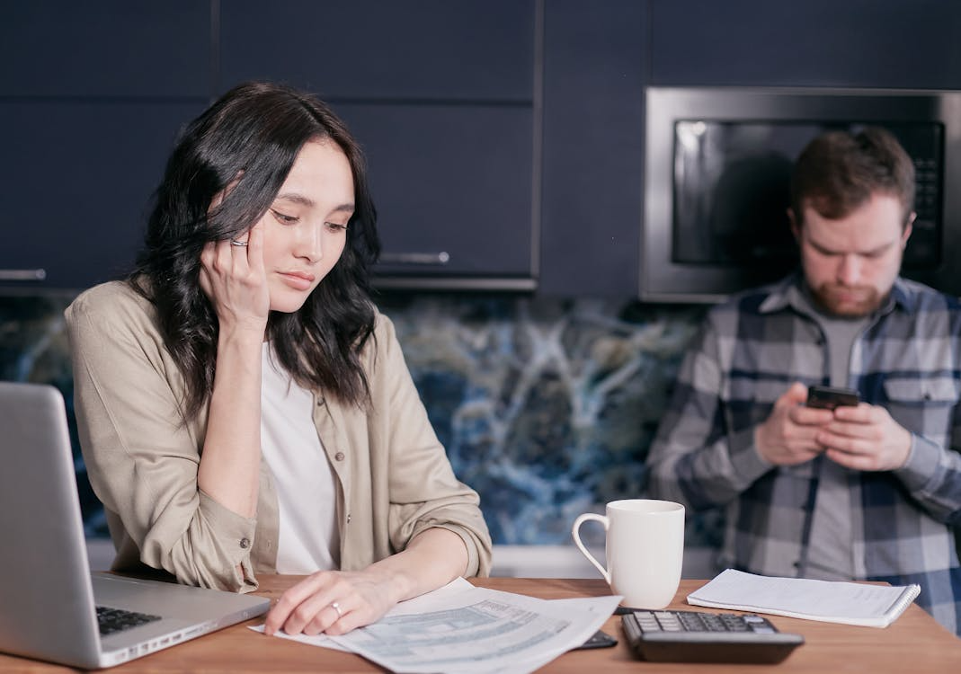 A woman studies paperwork at a kitchen table while a man in the background looks at his phone.