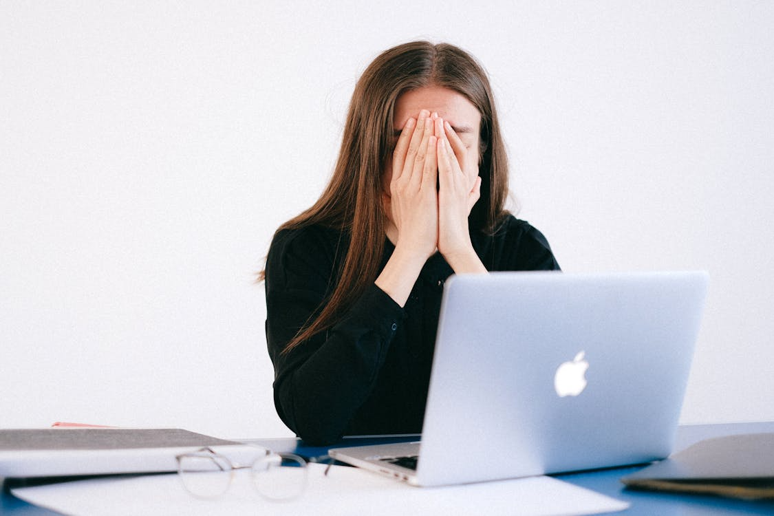 A person sitting at a desk with a laptop, covering their face with their hands, appears stressed or overwhelmed.