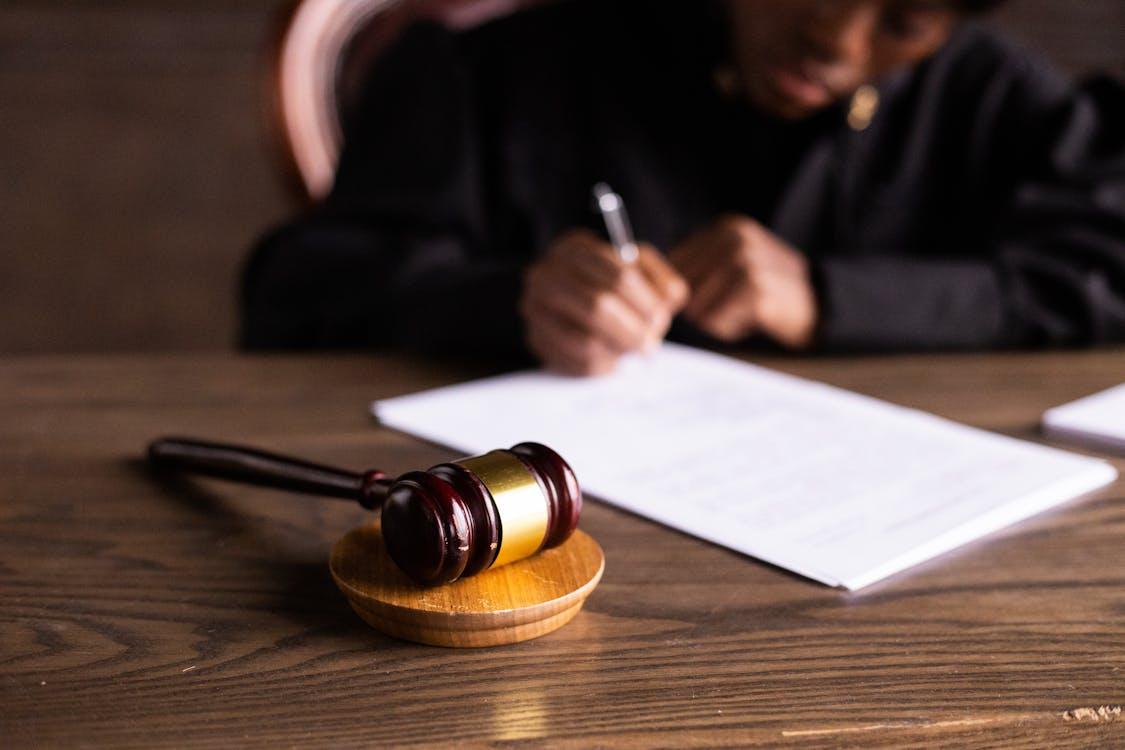 A judge’s gavel rests on a desk in the foreground while a person signs a legal document in the background.