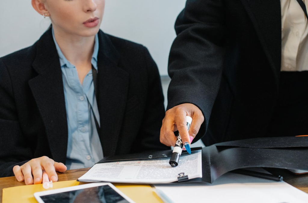 Two professionals review paperwork at a desk, with one person pointing to a document using a marker.