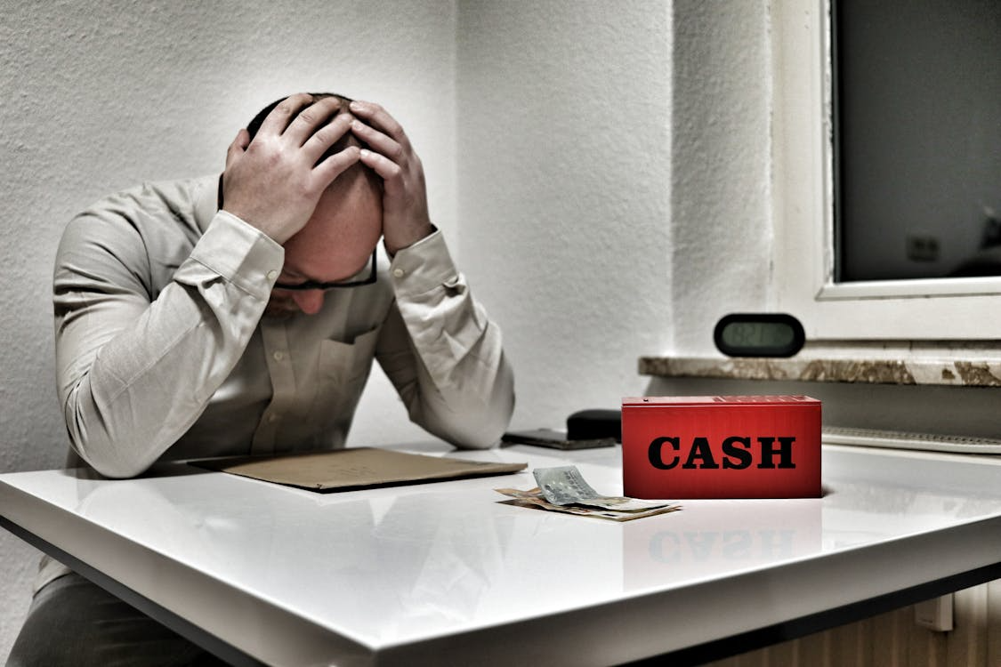 a stressed-out man sitting at a table with a red box saying cash placed in front of him