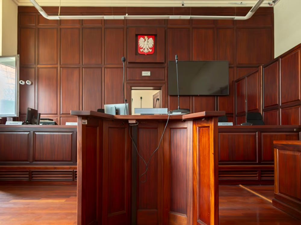 Empty courtroom with wood-paneled walls, a central witness stand with microphones and judges’ benches behind it