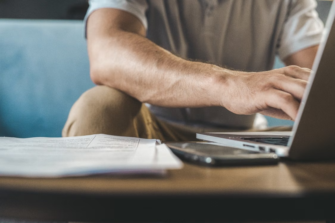 Person typing on a laptop beside a stack of papers and a phone on a wooden table